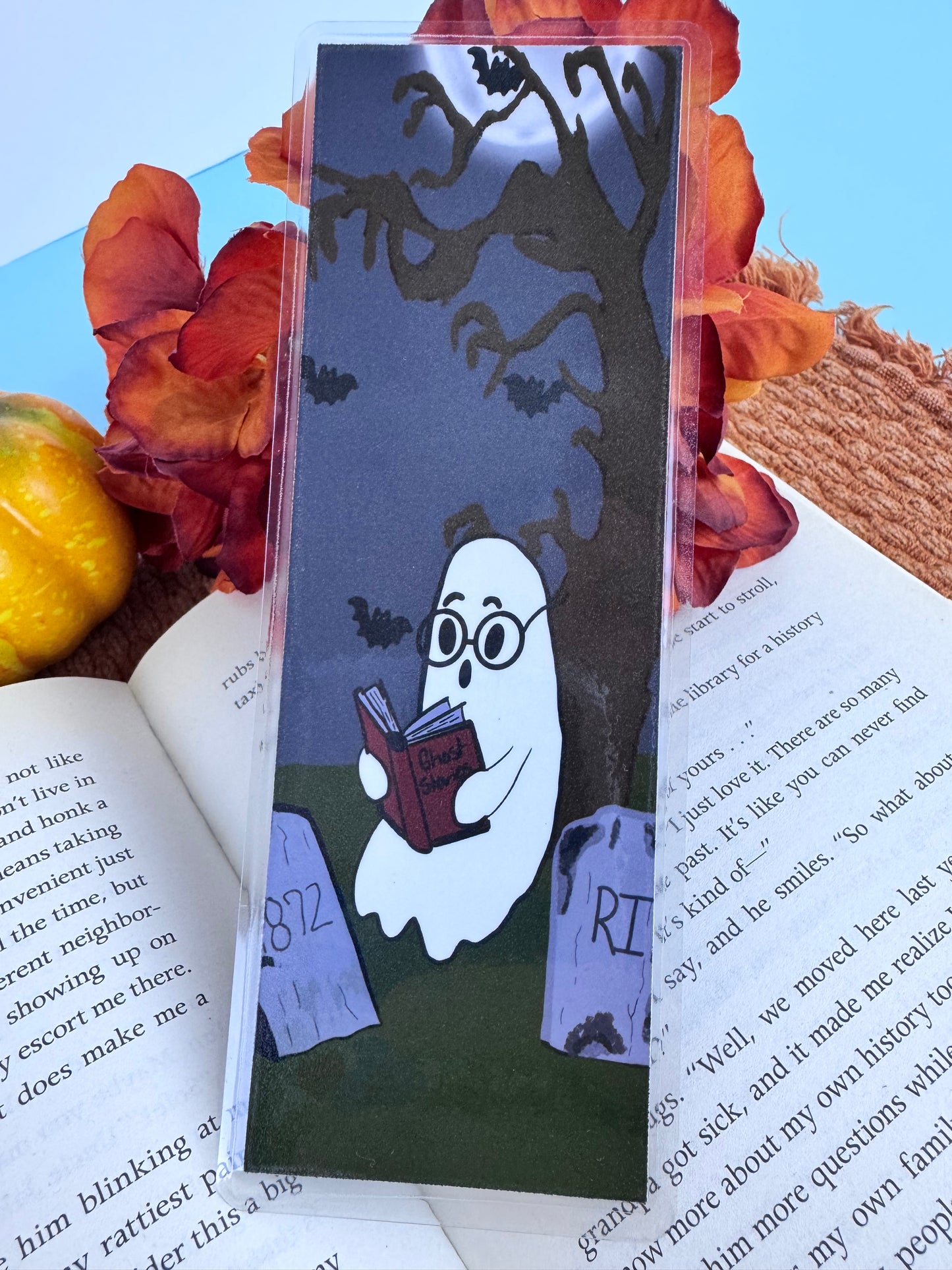 Handmade laminated bookmark featuring a cute ghost sitting in a cemetery under a spooky tree reading a book of ghost stories to bats.