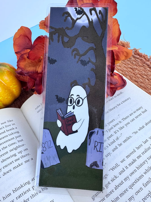 Handmade laminated bookmark featuring a cute ghost sitting in a cemetery under a spooky tree reading a book of ghost stories to bats.